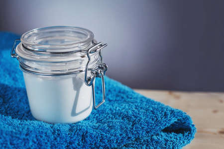 Transparent glass can filled with white body cream placed on top of bright blue folded towel on light wooden surfaceの写真素材