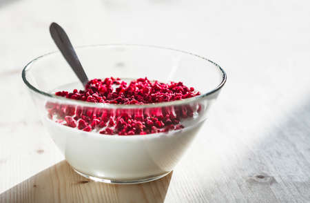 Transparent simple glass bowl filled with plain white yogurt topped with freeze dried raspberries placed on a light wooden surfaceの写真素材