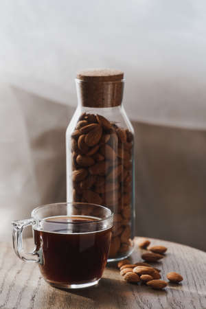 Small transparent cup of coffee and tall glass bottle filled with nuts surrounded by almonds on a...