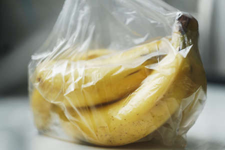 A bunch of almost ripe bananas wrapped in a transparent plastic bag on white table surfaceの写真素材