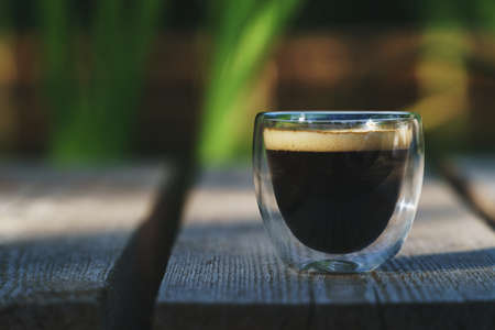 Small transparent double wall glass cup of fresh brewed coffee placed on a natural wooden surface in...