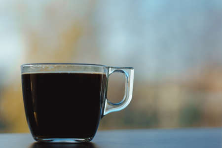 Small transparent cup of black coffee. Placed on dark wooden table in front of window. Blurred autumn sky and yellow trees in background.の写真素材