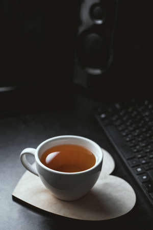 Blurry background image of white cup of tea and wooden heart-shaped plate on black office deskの写真素材