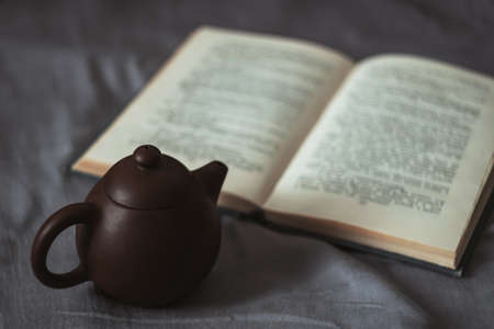 Dark soft picture of small brown clay teapot and open paper book on gray cloth covered surfaceの写真素材