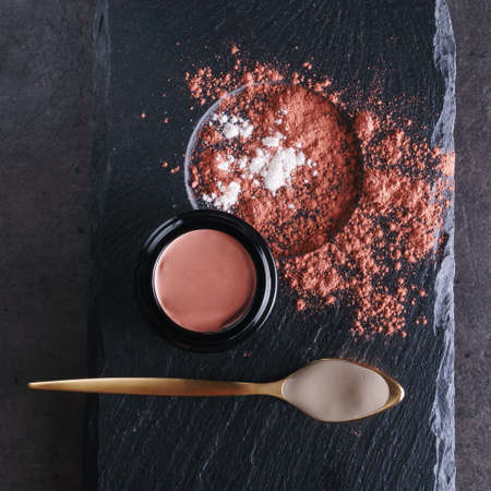 Beautiful photo of pink clay skincare product in open elegant black glass jar near golden spoon filled with creamy white clay surrounded by dry clay sprinkles on black stone plate surfaceの写真素材