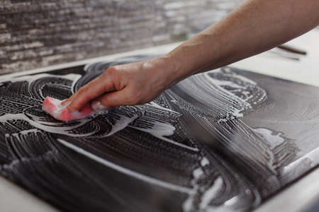 Hand cleaning black ceramic cooker surface with pink mop and foaming liquid