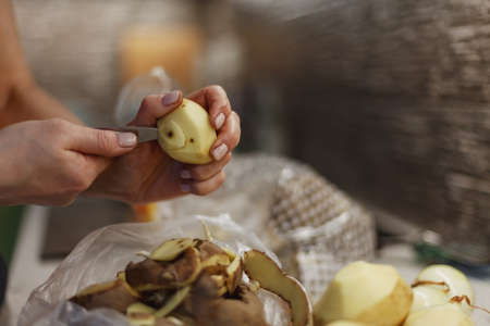 Woman's hands in process of peeling unwashed potato above small plastic bag