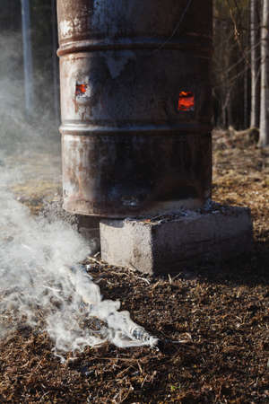 Fire safety - one smoking piece of firewood fallen out of old rusty metallic barrel full of burning...