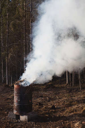 Heavy smoke flowing from rusty old metallic barrel while burning tree branches left after preparing...