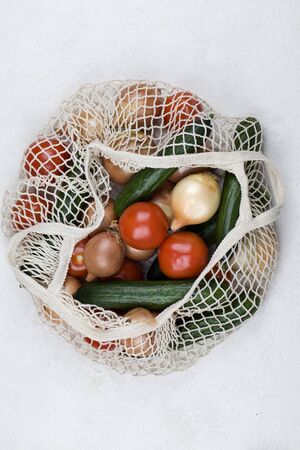 White mesh shopping bag with red tomatoes, bow and green cucumbers on white background. Top view,...