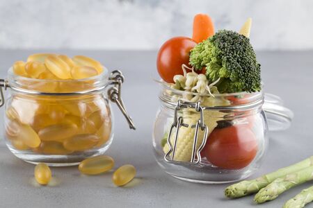 Yellow capsules in glass jar and fresh tasty vegetables on blue background. Nutrition multivitamins...