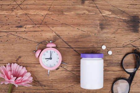 women health concept. medicine, health care style. copyspace. white plastic bottle with pills, flowers, glasses and clock on wooden background.の写真素材