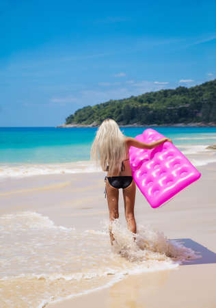 Slim blonde woman with pink swimming mattress on tropical beach outdoorsの写真素材