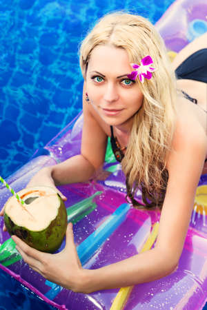 Young pretty blonde woman on air mattress in swimming pool with coconut cocktail drinkの写真素材