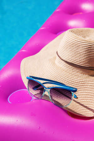 Sunglasses and straw hat on a pink air mattress in swimming pool. Tropical summer concept.