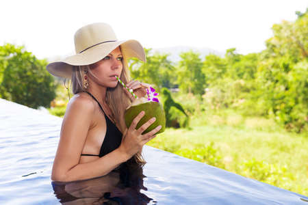 Young slim blonde woman in hat drinks healthy detox coconut juice in the swimming poolの写真素材