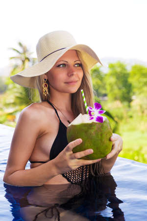 Young slim blonde woman in hat drinks healthy detox coconut juice in the swimming poolの写真素材