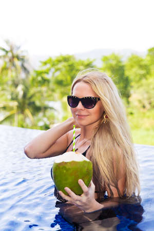Young slim blonde woman in sunglasses drinks healthy detox coconut juice in the swimming poolの写真素材