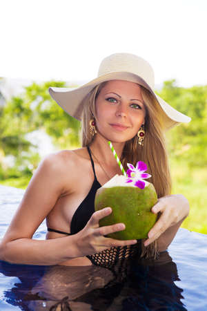 Young slim blonde woman in hat drinks healthy detox coconut juice in the swimming poolの写真素材