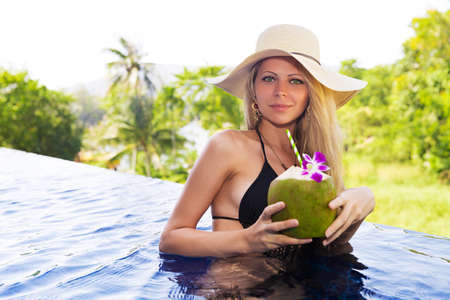 Young slim blonde woman in hat drinks healthy detox coconut juice in the swimming poolの写真素材