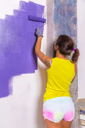 young attractive woman paints white wall and purple paint rollerの写真素材