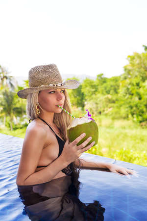 Young slim blonde woman in hat drinks healthy detox coconut juice in the swimming poolの写真素材
