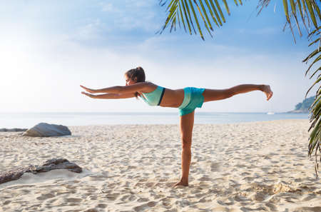 Young pretty slim brunette woman practise yoga Warrior on foot Virabhadrasana straight pose on a tropical beachの写真素材