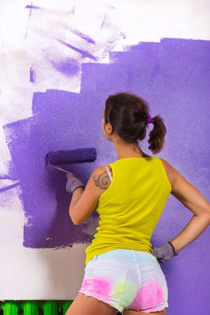 young attractive woman paints white wall and purple paint rollerの写真素材