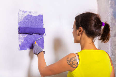 young attractive woman paints white wall and purple paint rollerの写真素材