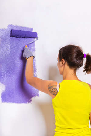 young attractive woman paints white wall and purple paint rollerの写真素材