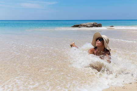 Young pretty slim brunette woman in hat and sunglasses swimming on the tropical beach in bikiniの写真素材