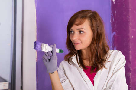 young attractive woman paints white wall and purple paint brushの写真素材
