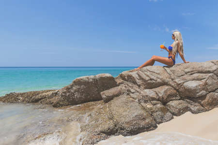 Slim blonde long haired woman in bikini with orange cocktail on tropical beachの写真素材