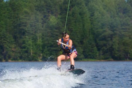 Young pretty slim brunette woman riding wakeboard on wave of motorboat in a summer lakeの写真素材