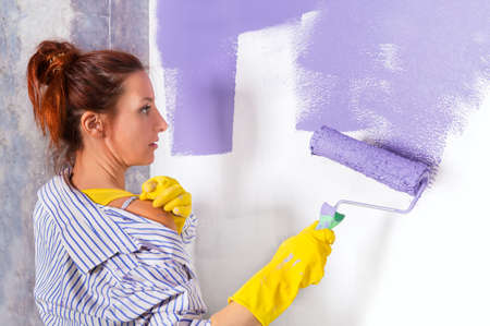 young attractive woman paints white wall and purple paint rollerの写真素材