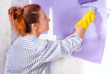 young attractive woman paints white wall and purple paint rollerの写真素材