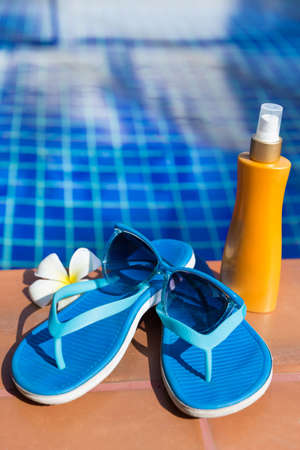 Blue slippers with sunscreen cream and sunglasses on border of a swimming pool - holiday tropical...