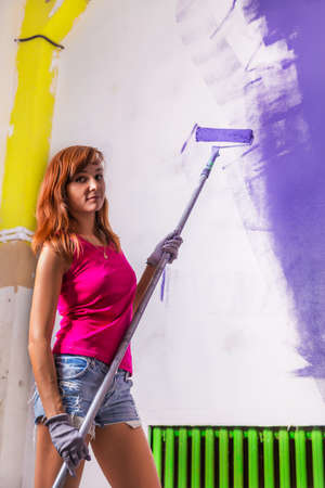 young attractive woman paints white wall and purple paint rollerの写真素材