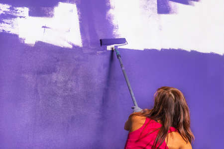 young attractive woman paints white wall and purple paint rollerの写真素材