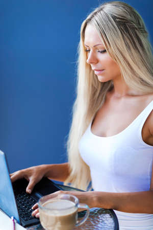 Young slim blonde woman freelancer with long white hair drinks coffee and works online with laptop in summer timeの写真素材