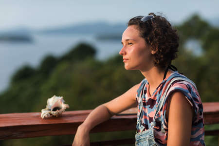 Unplugged curly haired woman sits on a tropical viewpoint observation deck and watches the landscape