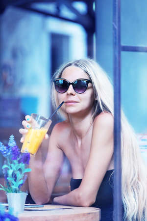 Young slim blonde woman with long hair drinks mango yellow smoothie fresh juice drink in cafe through glass windowの写真素材