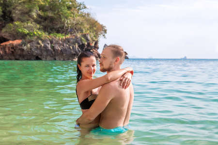 Young slim happy hipster couple of woman and man on a popular tropical Phra Nang Cave beach in Krabi, Thailand, in blue clear water kiss each otherの写真素材