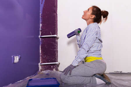 young attractive woman paints white wall and purple paint rollerの写真素材