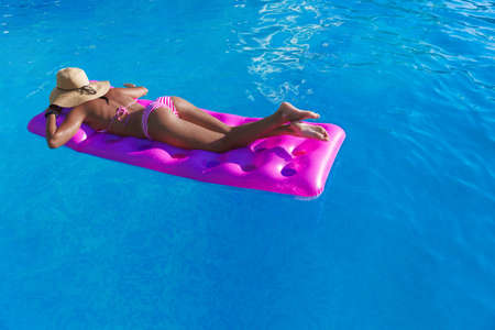 Young slim brunette woman in straw hat gets suntan on an air mattress in the swimming poolの写真素材