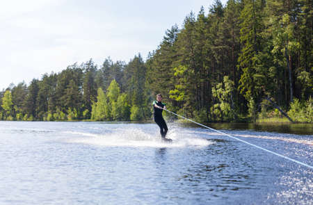 Young pretty slim brunette woman in wetsuit riding wakeboard on wave of motorboat in a summer lakeの写真素材