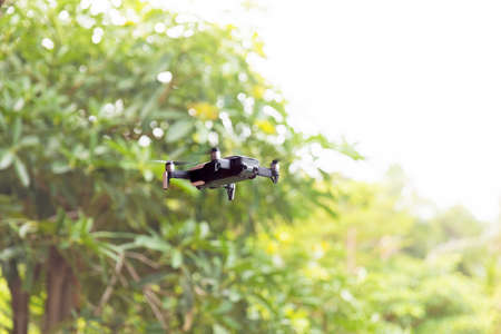 Small grey drone quadcopter flying near green trees daytime
