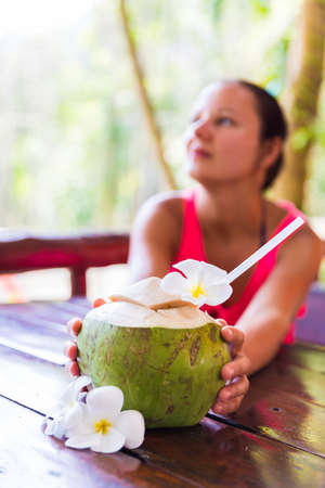 Fresh coconut cuts with tropical palm leaves and white frangipani flowers in front of woman in...