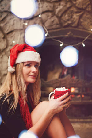 Pretty blonde woman near fireplace with cup of coffee with marshmallow in Christmas hatの写真素材