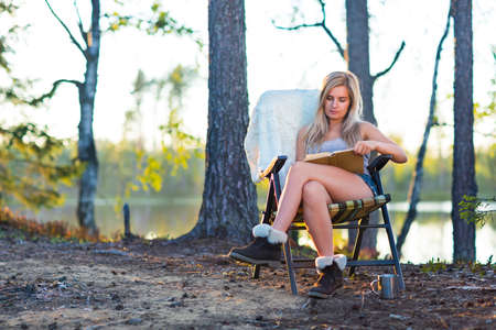 Young blonde woman in camping near lake reading a bookの写真素材
